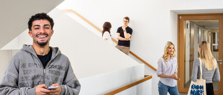 Ein Student steht im Treppenhaus des B6 Gebäudes. Er hat dunkle Haare, trägt einen grauen Hoodie und lacht. Er hat ein Handy in der Hand. 