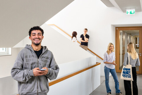 Ein Student steht im Treppenhaus des B6 Gebäudes. Er hat dunkle Haare, trägt einen grauen Hoodie und lacht. Er hat ein Handy in der Hand. 