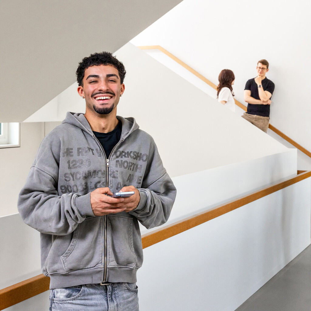 Ein Student steht im Treppenhaus des B6 Gebäudes. Er hat dunkle Haare, trägt einen grauen Hoodie und lacht. Er hat ein Handy in der Hand. 