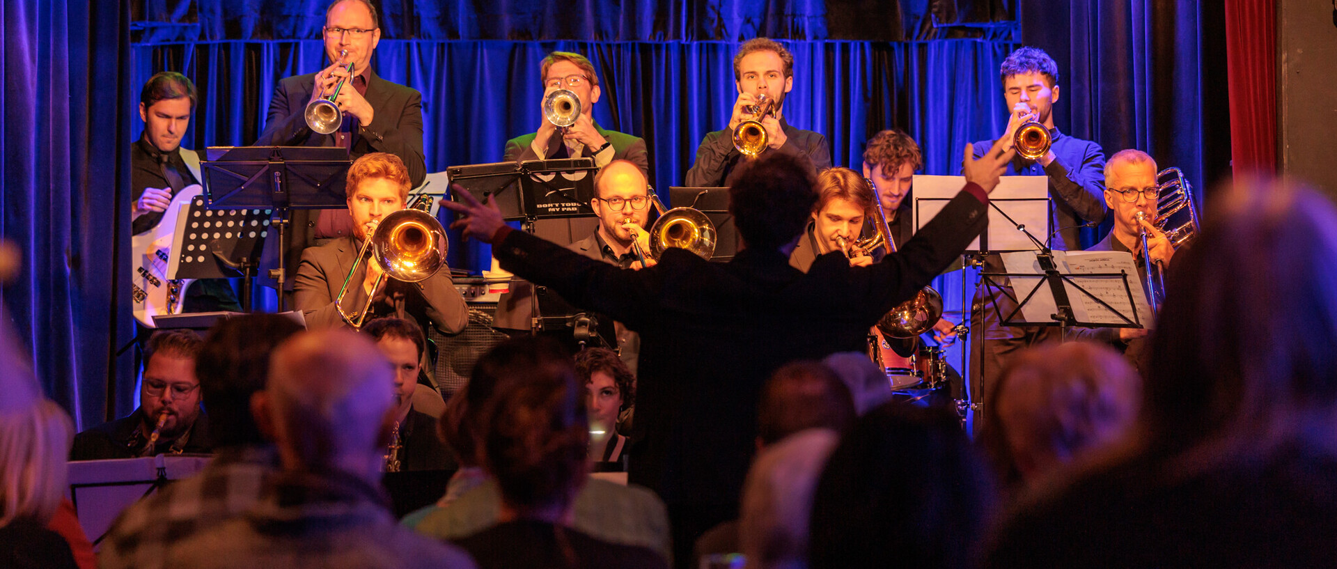 Eine Bigband spielt auf einer Bühne.Vor ihnen ist der Dirigent von hinten zu sehen, im Vordergrund sitzt Publikum.