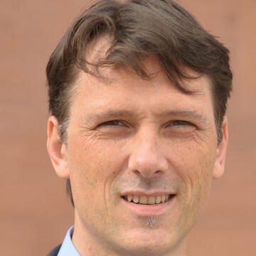 Prof. Dr. Thomas Bräuninger. Er trägt ein blaues Hemd und lächelt vor neutralem Hintergrund.