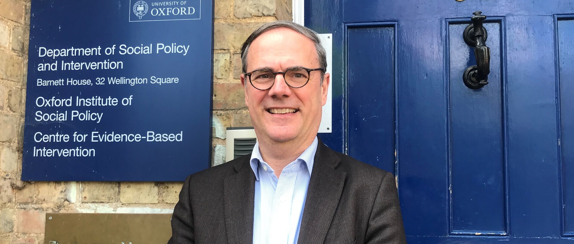 Prof. Dr. Ebbinghaus trägt ein hellblaues Hemd und ein dunkles Sakko und steht vor der blauen Haustür des Barnett House, Zentrum für evidenzbasierte Intervention der Universität Oxford