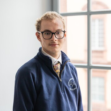 Paul trägt einen blauen Pullover über einem weißen Hemd und dazu eine Krawatte. Er hat kurze blonde Haare und eine Brille.