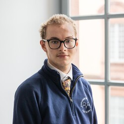 Paul trägt einen blauen Pullover über einem weißen Hemd und dazu eine Krawatte. Er hat kurze blonde Haare und eine Brille.