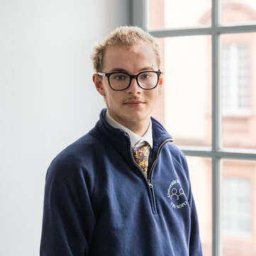 Paul is wearing a blue sweatshirt over a white shirt with a tie. He has short blond hair and is wearing glasses.