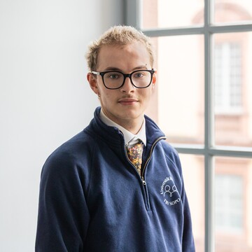 Paul trägt einen blauen Pullover über einem weißen Hemd und dazu eine Krawatte. Er hat kurze blonde Haare und eine Brille.