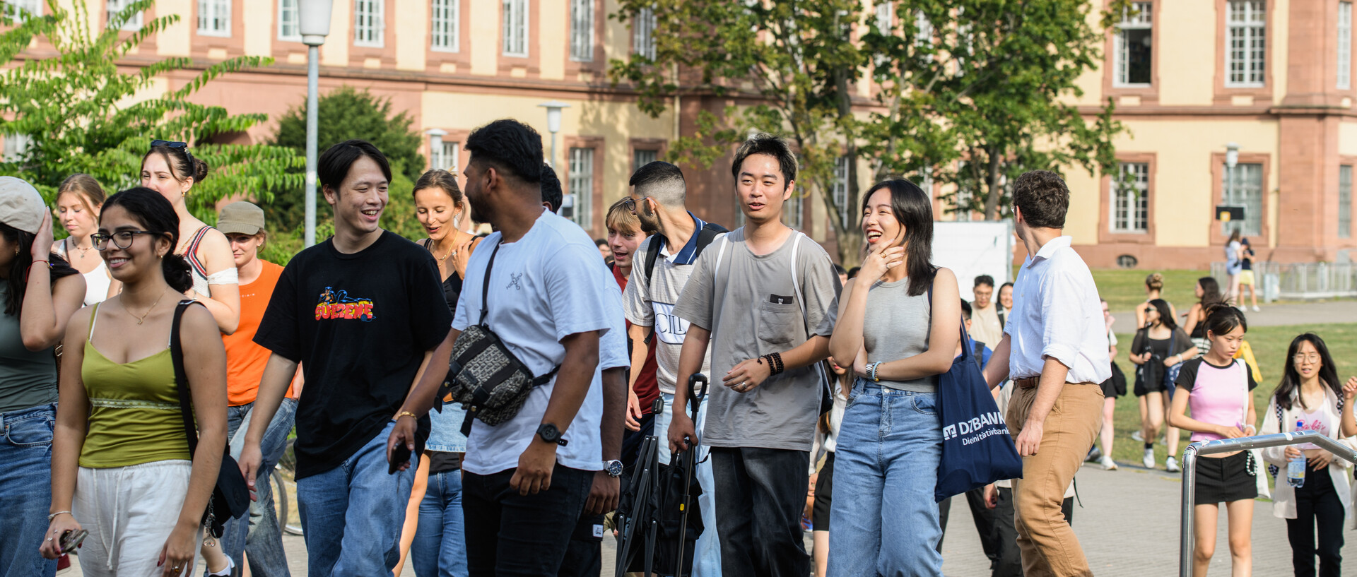 A group of students are chatting and walking across the lawn surrounding the Mensa. The Schloss is in the background.