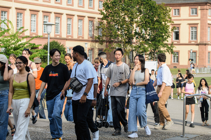 A group of students are chatting and walking across the lawn surrounding the Mensa. The Schloss is in the background.
