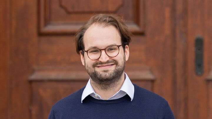 Portrait of Timo Kreisel. He is wearing a white shirt with a dark blue sweater over it and is standing in front of an entrance to Mannheim Palace.