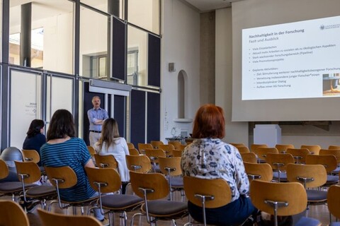 Vortrag im Schloss der Universität. Personen sitzen auf Stühlen und schauen nach vorne auf die Leinwand, auf der das Thema "Nachhaltigkeit in der Forschung" präsentiert wird.