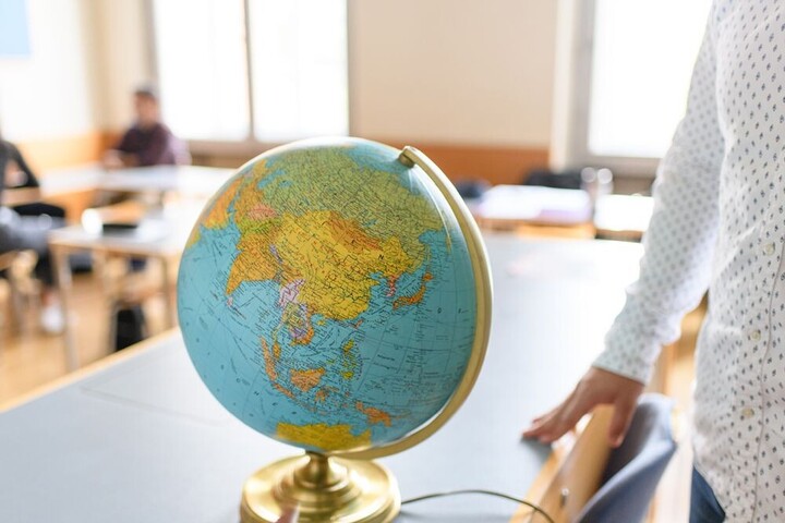 Ein Globus mit goldenem Ständer befindet sich auf einem Tisch. Im Hintergrund befinden sich weitere Tische und Stühle mit zwei verschommenen Personen. Auf der rechten Bildseite befindet sich eine weitere teilweise abgebildete Person. 