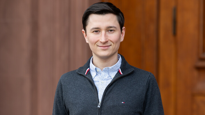 Christian hat kurzes braunes Haar und trägt ein blaues Hemd unter einer grauen Weste. Er lächelt und steht vor einer Holztür.
