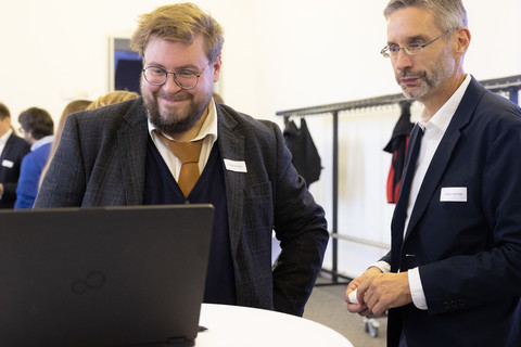 Zwei Männer schauen interessiert auf einen Laptop, der auf einem Bistro-Tisch steht.