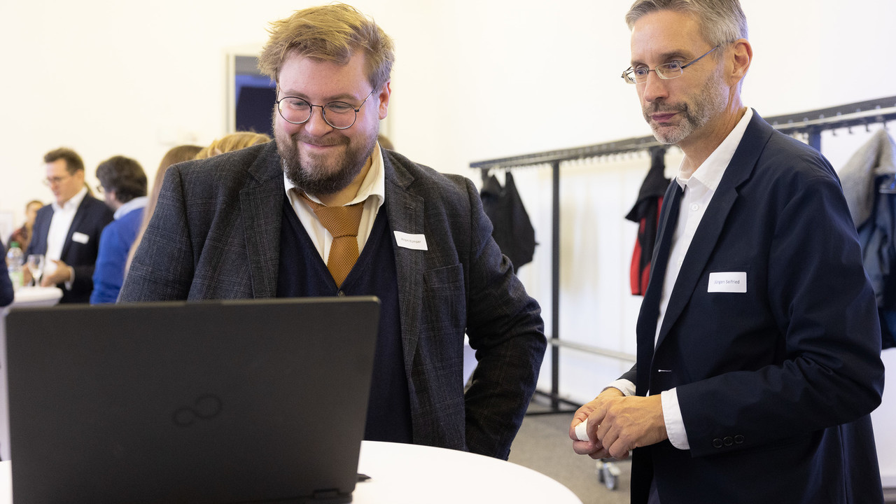 Zwei Männer schauen interessiert auf einen Laptop, der auf einem Bistro-Tisch steht.