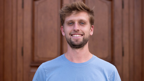 Timo Haupt. He is wearing a blue T-shirt and is standing in front of a brown door.