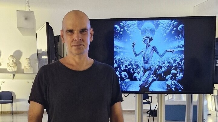 Timo Schuster trägt ein blaues T-Shirt und hat eine Glatze. Er steht vor einem Bildschirm, auf dem eine künstlich-maschinell aussehende Frau abgebildet ist.
