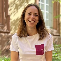 Anna Sattler sitzt lachend auf einer kleinen Steinmauer an der Uni. Sie trägt die braunen Haare offen und hat ein sommerliches Outfit an..