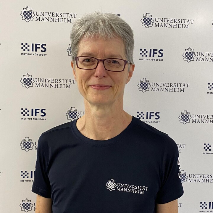 Bianka Velte hat kurze graue Haare. Sie trägt eine Brille und ein blaues T-Shirt mit der Aufschrift Universität Mannheim. Sie steht vor einer Stellwand, auf denen die Logos der Uni Mannheim und des IFS mehrfach abgebildet sind.