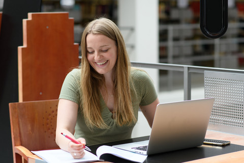 Eine Studierende sitzt lächelnd mit einem Laptop und Notizblock an einem Schreibtisch aus rotem Holz.