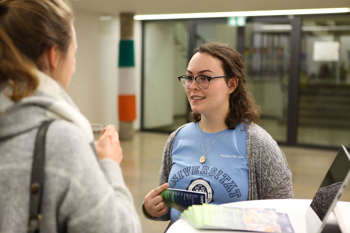 Zwei Studentinnen unterhalten sich. Eine Studentin hält einen Flyer in der Hand und trägt ein hellblaues T-Shirt des Instituts für Sport der Universität Mannheim.