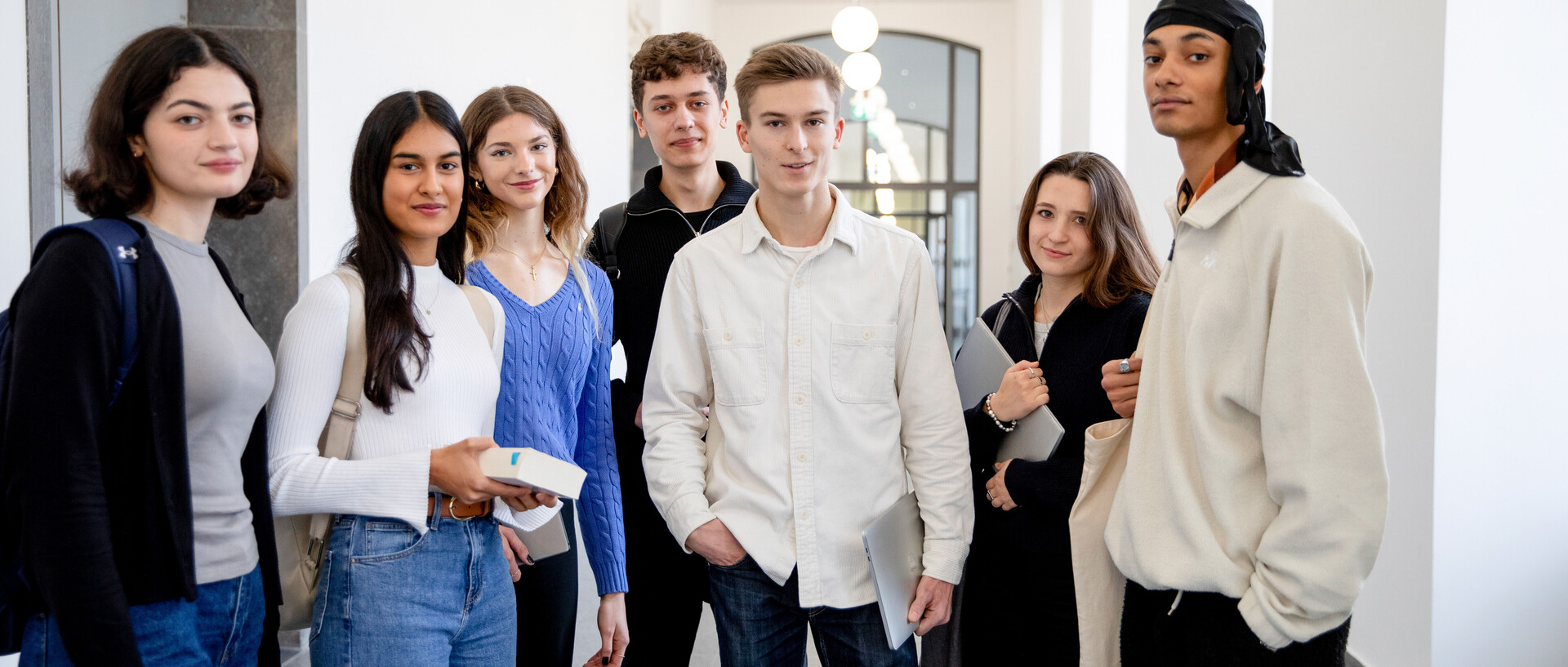 Eine Gruppe von Studierenden steht im Flur der Uni. Sie haben Bücher, Laptops und Rucksäcke dabei.