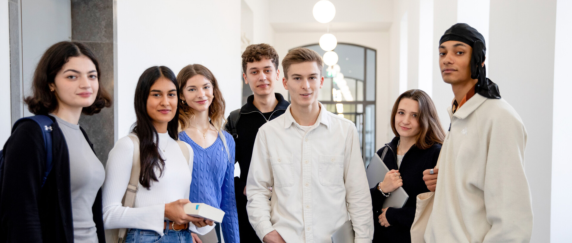 Eine Gruppe von Studierenden steht im Flur der Uni. Sie haben Bücher, Laptops und Rucksäcke dabei.