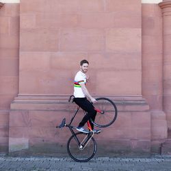 Der Kunstradfahrer Serafin Schefold hält das Vorderrad seines Fahrrads in die Luft und sitzt dabei auf dem Lenker. Er trägt ein weißes mit bunten Streifen.