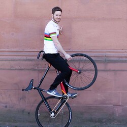 Artistic cyclist Serafin Schefold is holding the front wheel of his bicycle in the air while sitting on the handlebars. He is wearing a white shirt with colorful stripes.