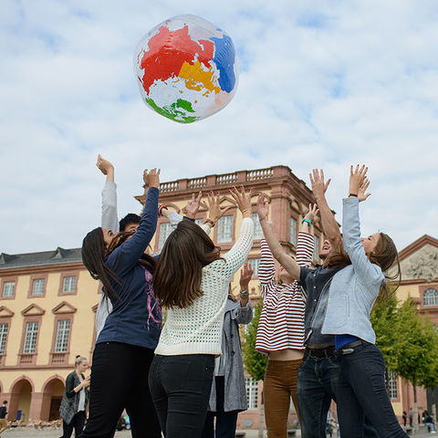 Eine Gruppe von sieben Studierenden steht in einem Kreis auf dem Ehrenhof. Sie werfen einen aufgeblasenen Globus hoch und haben die Hände in den Himmel gestreckt.