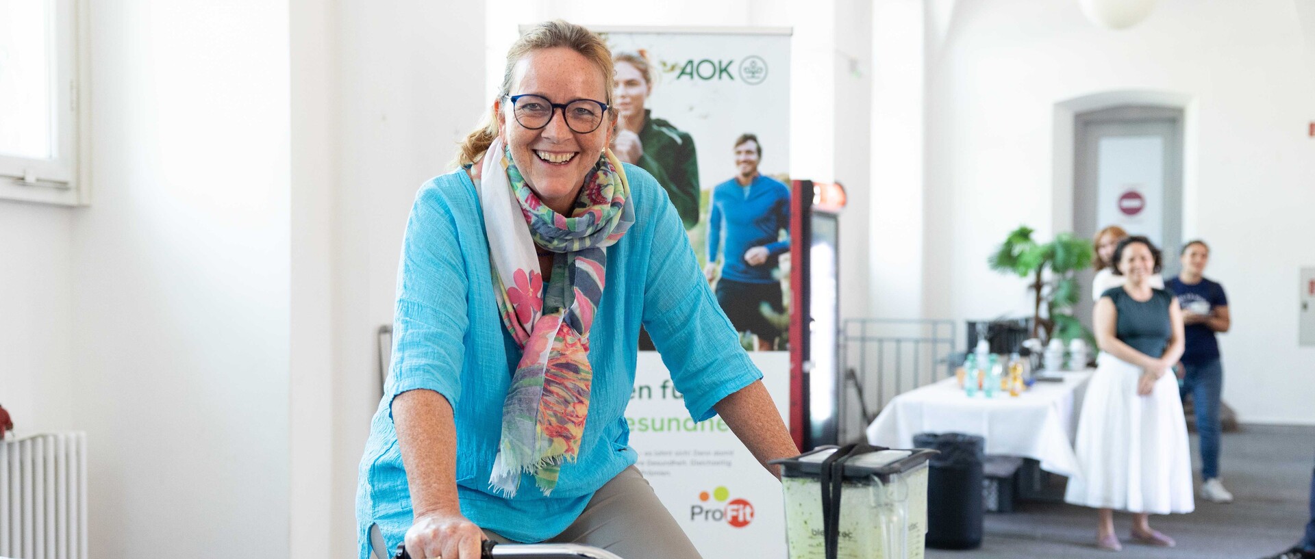 Eine Frau sitzt strahlend auf einem Ergometer, das an einen Mixer angeschlossen ist.