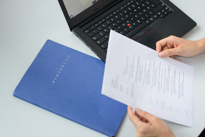 Eine blaue Bewerbungsmappe und ein schwarze Laptop liegen auf einem weißen Tisch. Eine Person hält einen Lebenslauf in den Händen (Blick über dei Schulter)