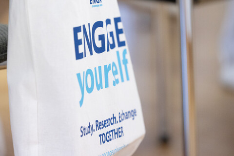 Eine Stofftascher hängt an einem Stuhl. Sie trägt die folgende Aufschrift: "Engage yourself. Study, research, exchange together."