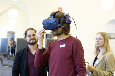 Ein junger Mann mit VR-Brille. Rechts und links von ihm eine Frau und ein Mann, die ihn lächelnd anschauen.