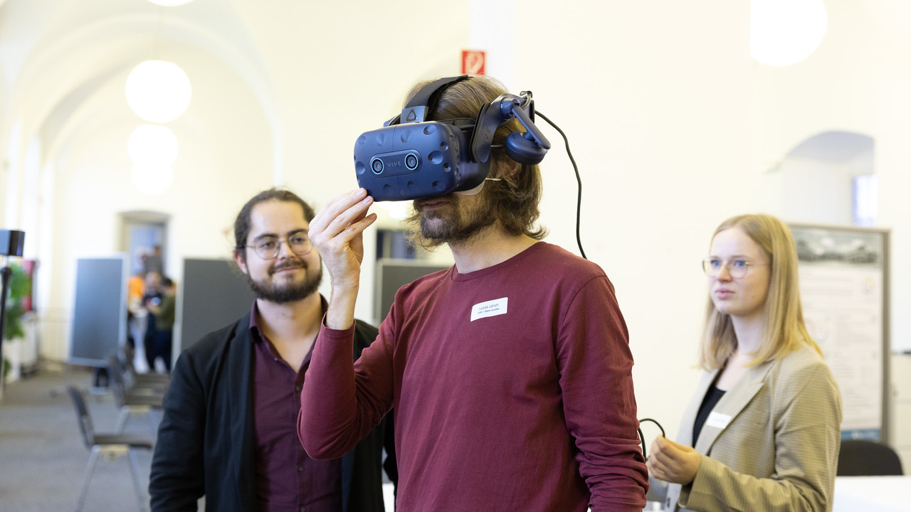 Ein junger Mann mit VR-Brille. Rechts und links von ihm eine Frau und ein Mann, die ihn lächelnd anschauen.