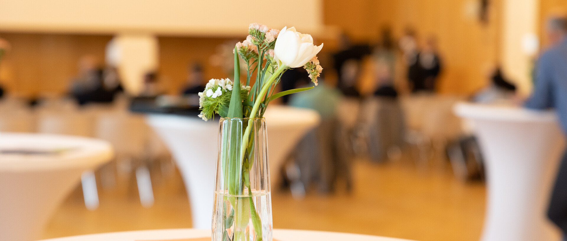 Blumenstrauß in einer Vase auf einem Tisch. Im Hintergrund weiße Stehtische und Personen.