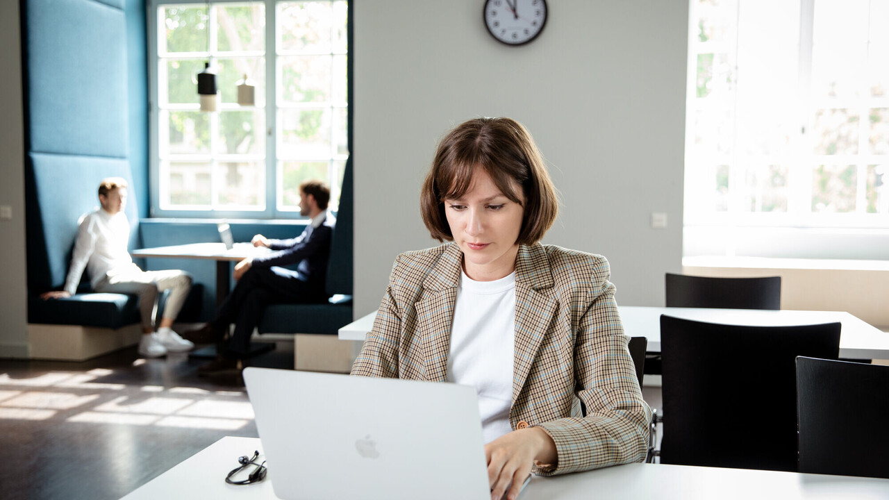 Eine Person sitzt mit Laptop an einem Tisch in der Bibliothek. Im Hintergrund sitzen zwei Studierende an einem Gruppentisch gegenüber und unterhalten sich.
