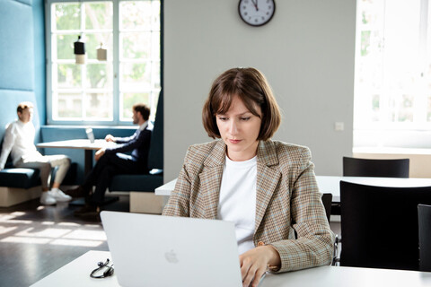 Eine Person sitzt mit Laptop an einem Tisch in der Bibliothek. Im Hintergrund sitzen zwei Studierende an einem Gruppentisch gegenüber und unterhalten sich. 
