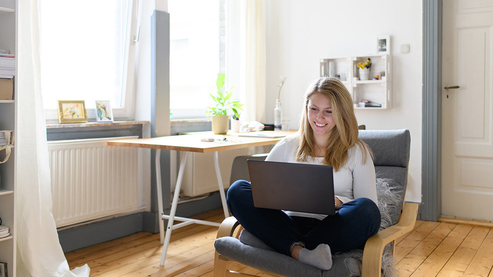 Eine junge Frau sitzt in einem gemütlichen Zimmer mit ihrem Laptop auf einem Sessel.