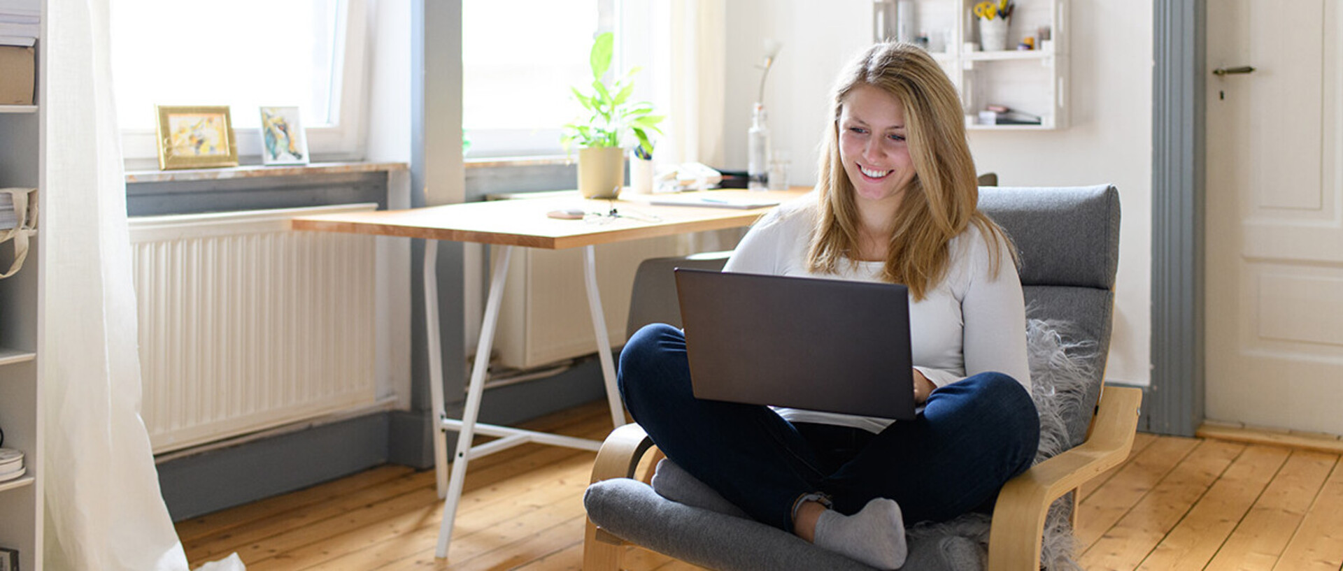 Eine junge Frau sitzt in einem gemütlichen Zimmer mit ihrem Laptop auf einem Sessel.