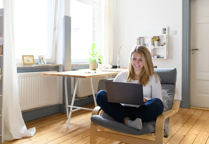 Eine junge Frau sitzt in einem gemütlichen Zimmer mit ihrem Laptop auf einem Sessel.