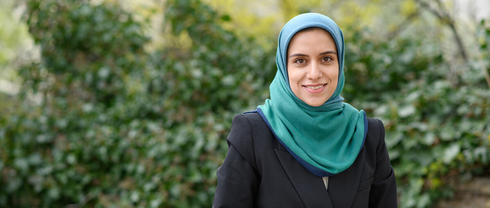 A young woman is standing smiling in front of a background filling green hedge.