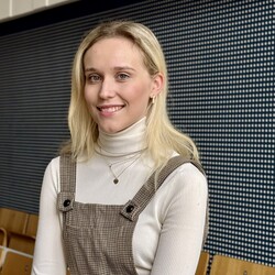 Porträt von Anna-Lena Baumann. Anna-Lena Baumann trägt eine braun-karrierte Latzhose, einen hellen Rolli und dezenten Goldschmuck. Sie hat blonde schulterlange Haare, die sie offen trägt. Sie lacht keck in die Kamera.