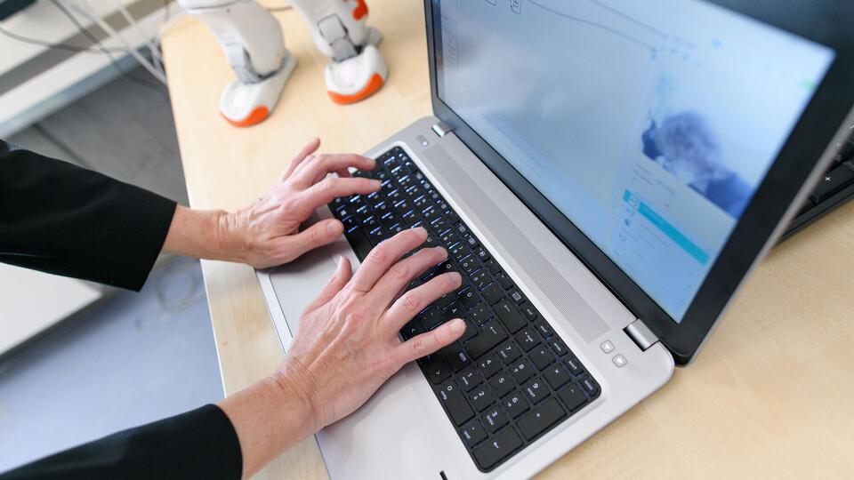 Eine Person tippt auf der Tastatur eines Laptops, neben dem auf dem Tisch ein Roboter steht.
