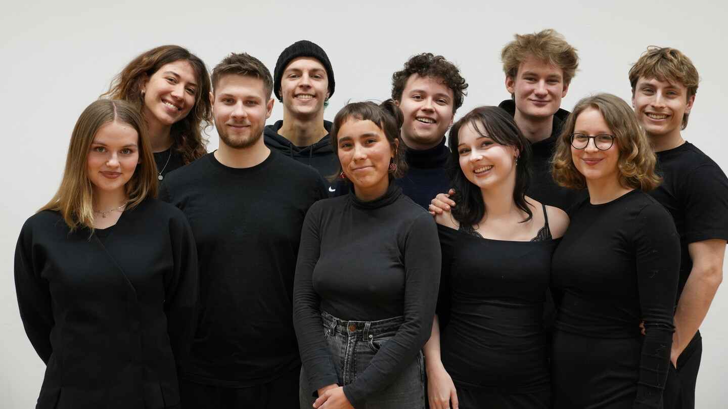 Zehn schwarz gekleidete Studierende stehen in zwei Reihen vor einem weißen Hintergrund und lächeln in die Kamera.