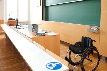 Ein Vorlesungssaal mit Tischen und einer Tafel. Unten im Bild ist ein Rollstuhl und davor ein Tisch mit einem Schild in blau-weiß auf dem ein Rollstuhl ist. Der Tisch ist hoch genug, dass ein Rollstuhl darunter fahren kann. 