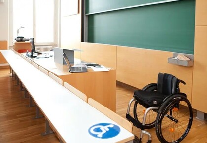Abgebildet ist ein Vorlesungssaal mit Tischen und einer Tafel. Unten im Bild ist ein Rollstuhl abgebildet und davor ein Tisch mit einem Schild in blau-weiß auf dem ein Rollstuhl ist. Der Tisch ist hoch genug, dass ein Rollstuhl darunter fahren kann.