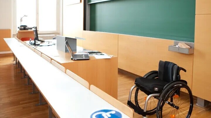 Ein Vorlesungssaal mit Tischen und einer Tafel. Unten im Bild ist ein Rollstuhl und davor ein Tisch mit einem Schild in blau-weiß auf dem ein Rollstuhl ist. Der Tisch ist hoch genug, dass ein Rollstuhl darunter fahren kann.