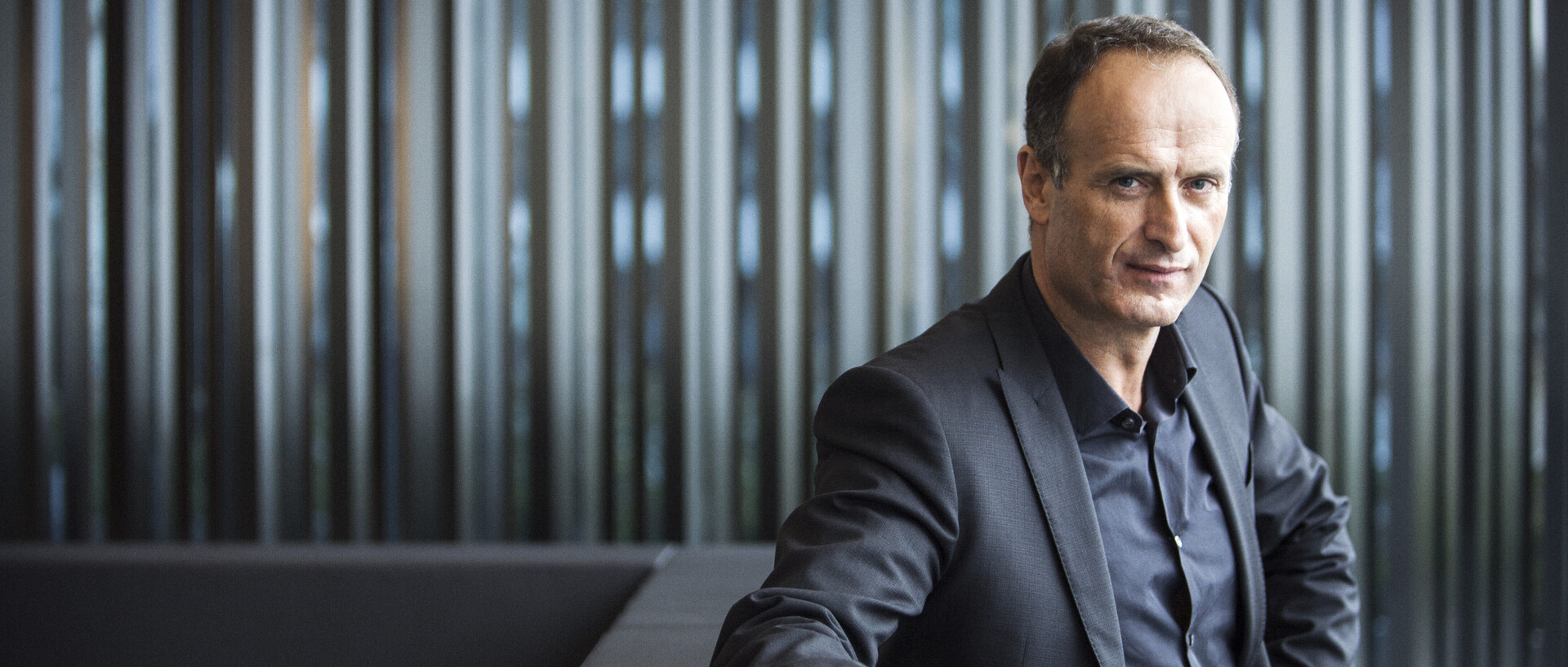 Dr. Bruno Sälzer sitzt in einem dunklen Anzug, einen Arm abgestützt, den anderen in der Seite, auf einer grauen Bank