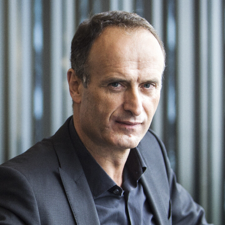 Dr. Bruno Sälzer sitzt in einem dunklen Anzug, einen Arm abgestützt, den anderen in der Seite, auf einer grauen Bank