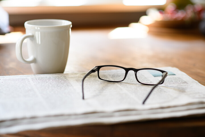 Eine Zeitung, auf der eine Lesebrille liegt. Daneben steht eine Kaffeetasse.